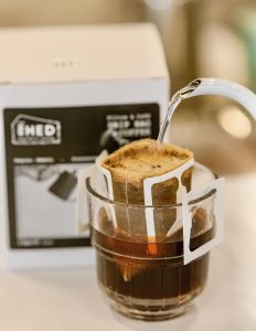 Hot water in a kettle is slowly poured over The Shed Coffee's drip bag specialty coffee product. The single-use drip bag coffee product has two elegant arms shown here propping the filter open over a glass. The glass is filling with coffee as the hot water filters through the product.