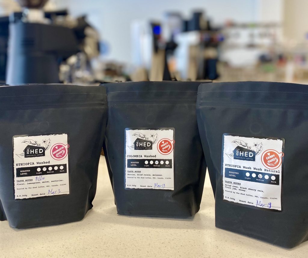 Three bags of The Shed Coffee's freshly roasted coffee stand together on a counter at a coffee bar. The bags are bag with labels on the front describing each coffee bean's variety, tasting notes, and origin. The labels feature the logo of The Shed Coffee.